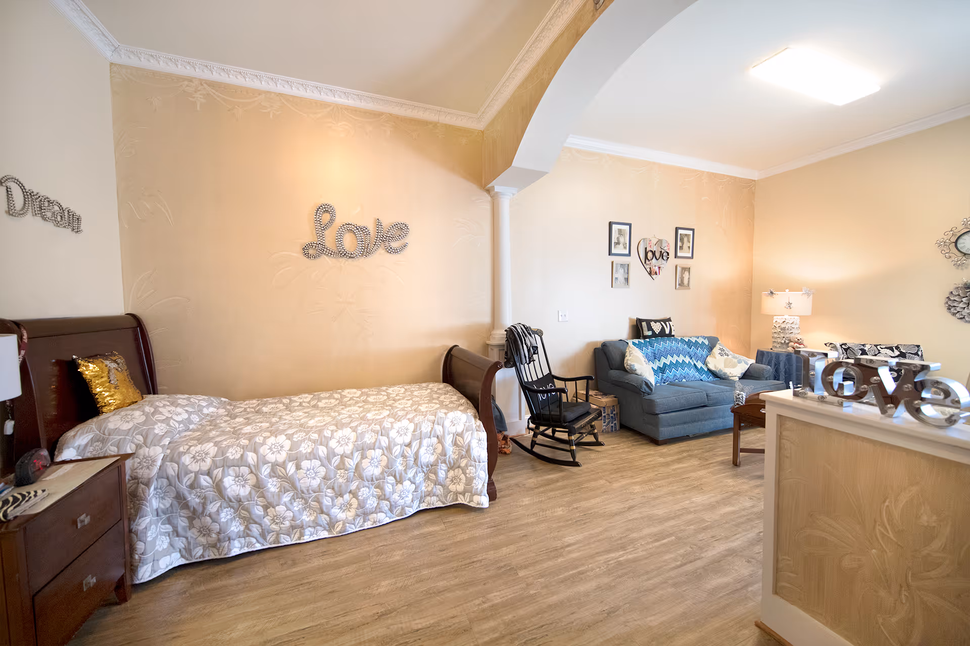 A cozy room with a single bed covered in a floral quilt, a wooden nightstand with a lamp, and a rocking chair. The walls are decorated with the words 'Love' and 'Dream' in decorative lettering. There is a blue loveseat with patterned pillows, a small wooden coffee table, and a lamp on a side table. The room has light-colored walls and wood flooring, with a decorative archway and column separating the bed area from the sitting area.