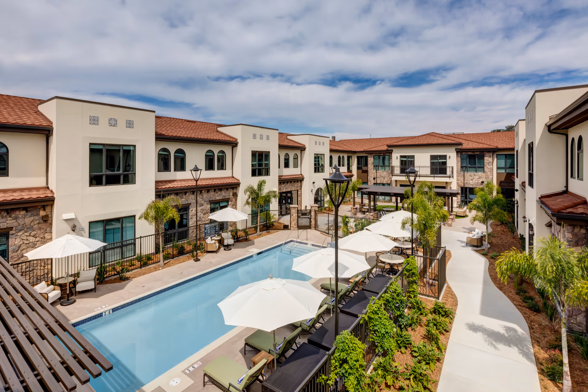 Outdoor courtyard with a rectangular swimming pool, lounge chairs and umbrellas flanked by two-story Mediterranean-style buildings.