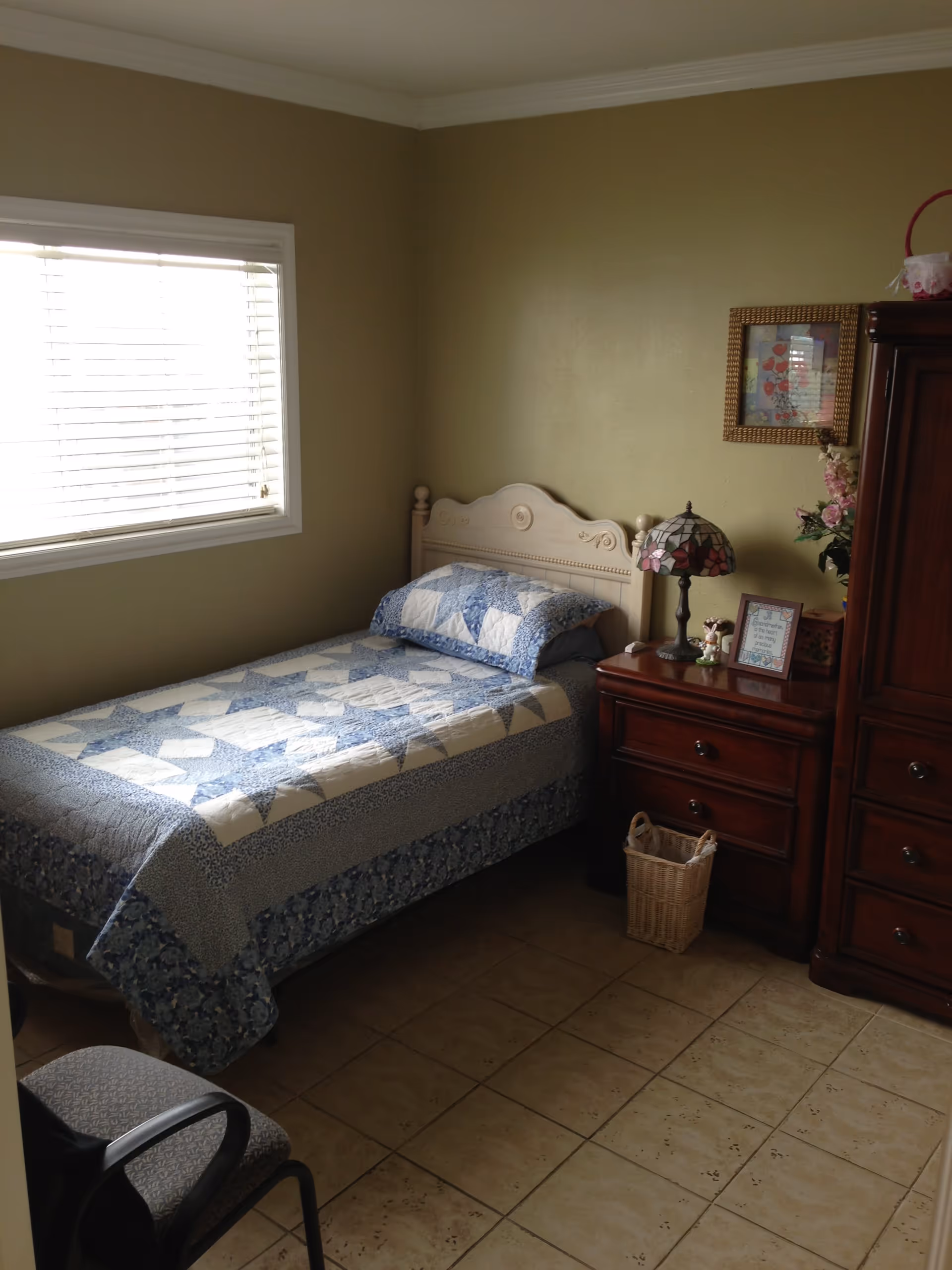 A small bedroom with a single bed covered with a blue and white quilt. Next to the bed is a wooden nightstand with a decorative lamp, a framed picture, and some flowers. There is a wooden wardrobe beside the nightstand and a window with blinds on the left wall. A chair is partially visible in the foreground.