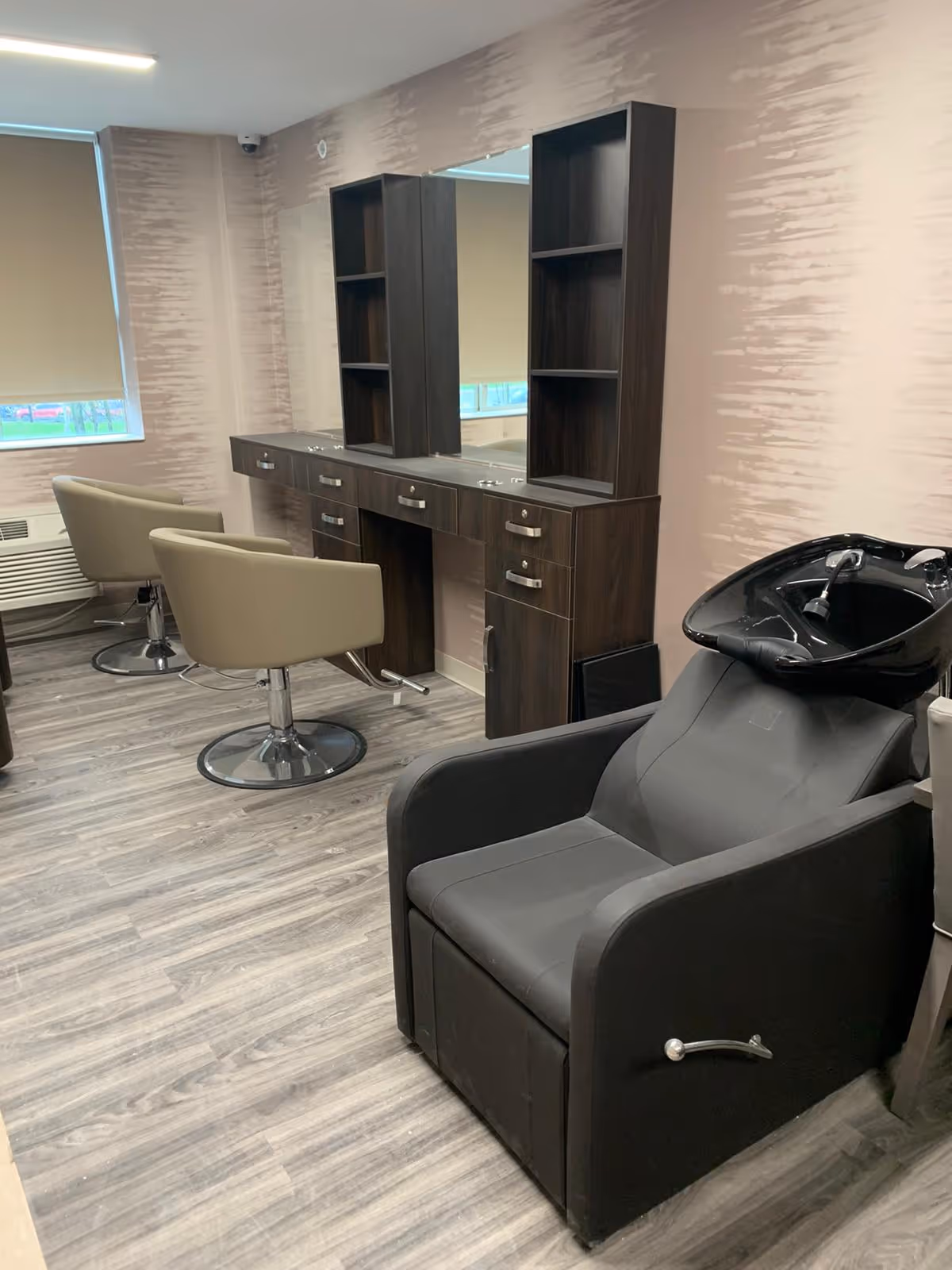Interior of a salon area with two beige swivel chairs in front of a long dark wood vanity with mirrors and shelves. In the foreground, there is a black shampoo chair with a wash basin. The room has light wood flooring and light pink textured walls with a window covered by a beige roller shade.