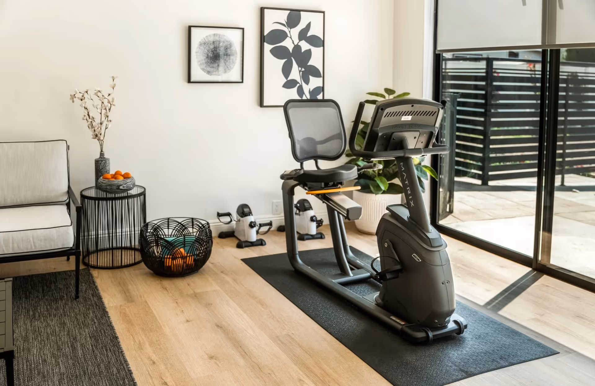 A modern exercise room with a recumbent stationary bike on a black mat near large sliding glass doors. The room has light wood flooring, a small black wire basket with oranges, a side table with a vase and more oranges, a cushioned chair, two framed abstract art pieces on the wall, and a potted plant in the corner.