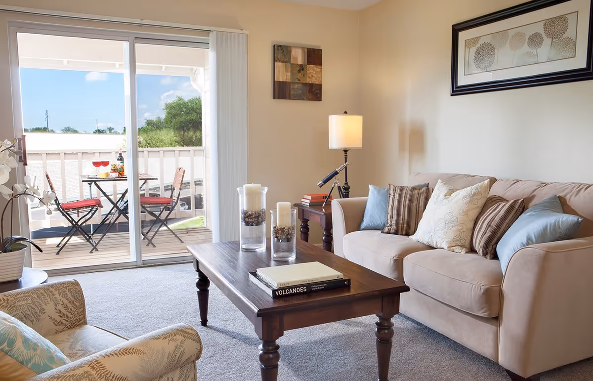 A cozy living room with a beige sofa adorned with multiple cushions, a wooden coffee table with candles and books, a side table with a lamp and telescope, and a sliding glass door leading to a balcony with a small table and two chairs with red cushions.