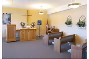 Small indoor chapel with wooden pews facing a pulpit and a wall-mounted cross, floral arrangements and an American flag.