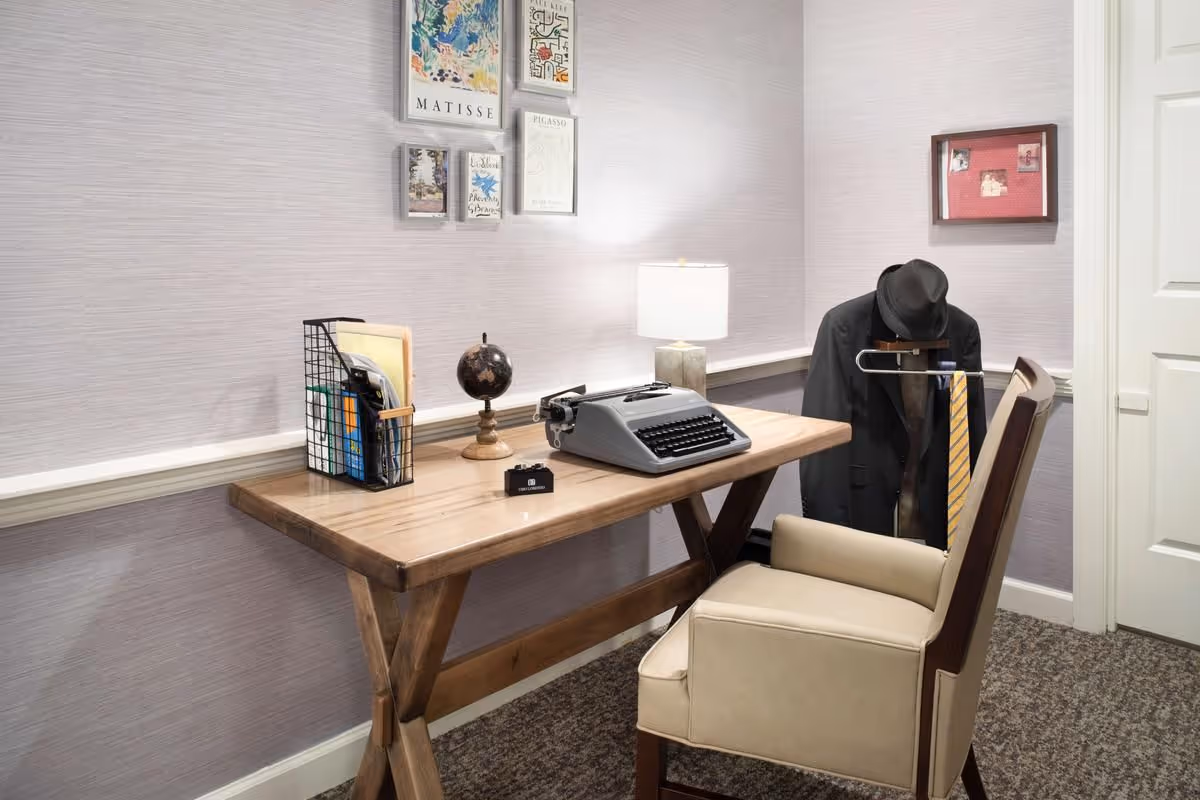 A small office or study area with a wooden desk holding a vintage typewriter, a small globe, a desk lamp, and a wire organizer with folders and papers. A beige upholstered chair is positioned in front of the desk. On the right side, a coat rack holds a black jacket, a black hat, and a yellow striped tie. The walls are light purple with framed artwork and photos hanging above the desk and on the adjacent wall.