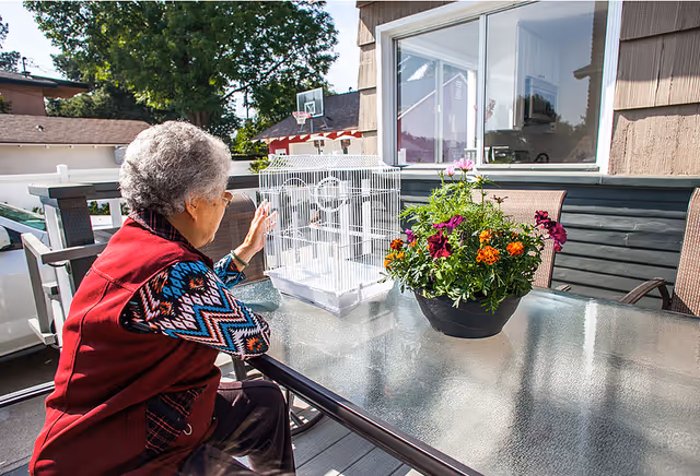 An elderly woman with gray hair and glasses sits at a glass outdoor table on a patio. She is wearing a red vest over a patterned long-sleeve shirt and is reaching towards a white birdcage on the table. Next to the birdcage is a black pot with colorful flowers. In the background, there is a basketball hoop and part of a house with windows.