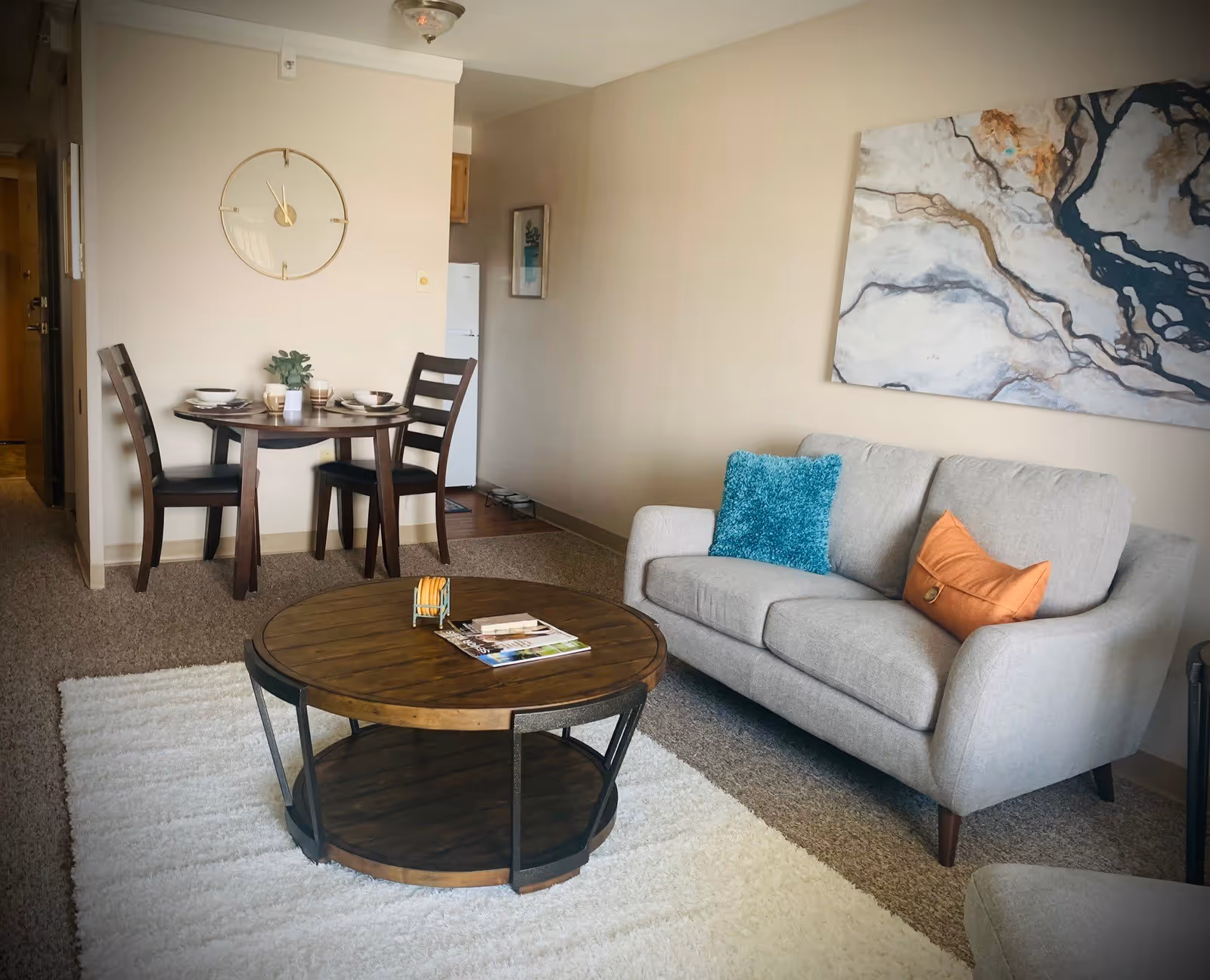 A cozy living room area in an assisted living center featuring a light gray sofa with blue and orange throw pillows, a round wooden coffee table on a white rug, and a small dining table with two chairs set against a beige wall with a large round clock. A piece of abstract artwork hangs above the sofa, and a kitchen area with a refrigerator is visible in the background.