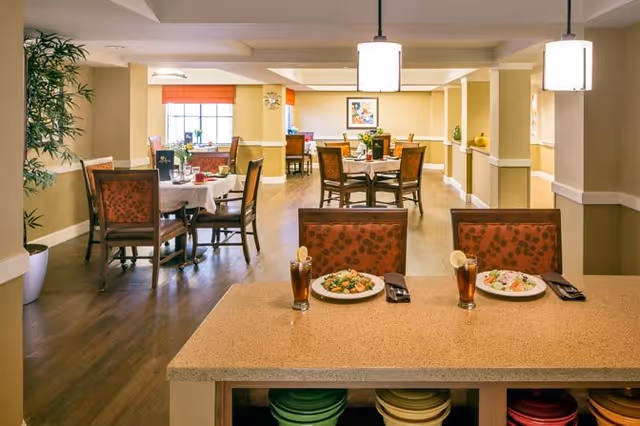 A bright dining room in a senior living facility with several tables and chairs arranged neatly. Two plates of food and two glasses of iced tea with lemon slices are placed on a countertop in the foreground. The room has wooden flooring, beige walls, pendant lights, and a large window with orange blinds in the background. There are decorative plants and framed artwork on the walls.