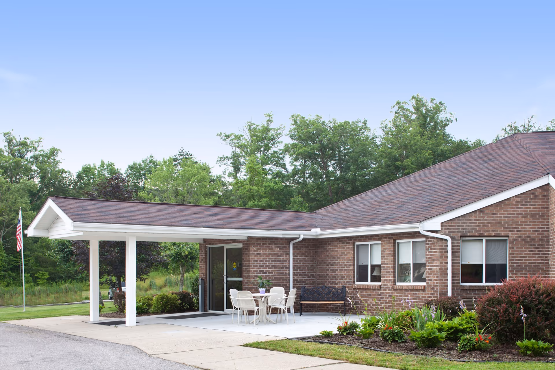 Exterior view of a single-story brick building with a covered entrance supported by white columns. There is a small patio area with a table and chairs, a bench, and landscaping with green plants and flowers. An American flag is visible on a flagpole to the left, and trees are in the background under a clear blue sky.