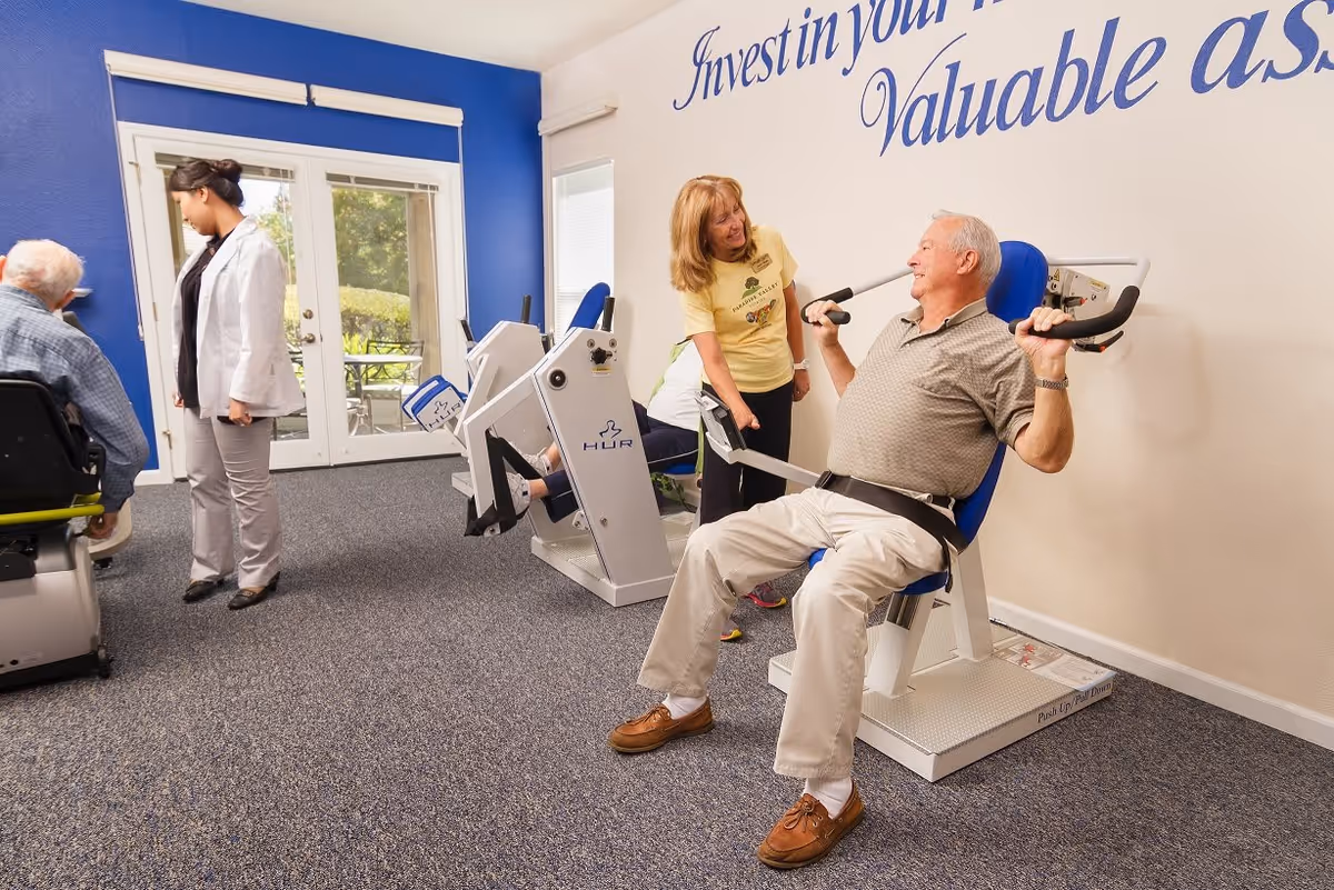 An elderly man exercises on a seated chest press machine in a fitness room while a woman in a yellow Paradise Valley Estates t-shirt encourages him. Another elderly man is seated on a different exercise machine, and a woman in a white coat stands nearby. The room has blue and white walls, carpeted floor, and large glass doors leading outside. Text on the wall reads 'Invest in your valuable ass...'.