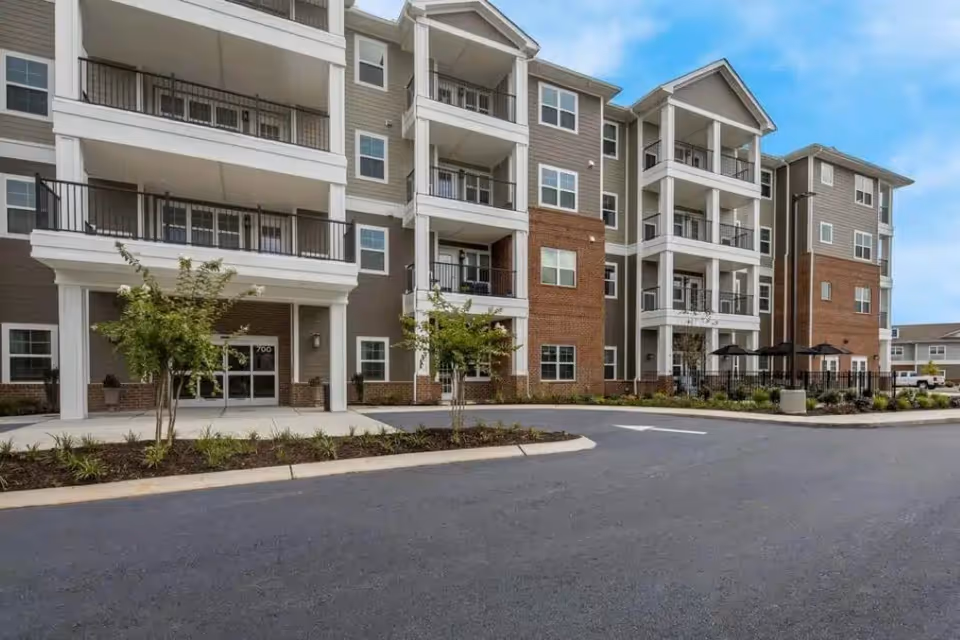 Exterior view of a multi-story senior living facility building with balconies, windows, and a driveway in front. There are small trees and landscaped areas near the entrance.