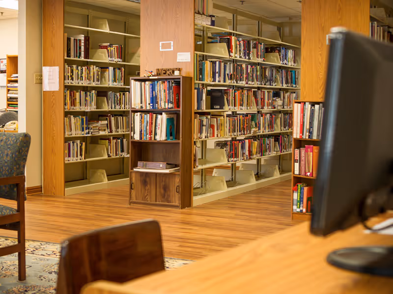 Interior view of a library area in a senior living facility with wooden bookshelves filled with books, a wooden floor, and a computer monitor on a desk in the foreground.
