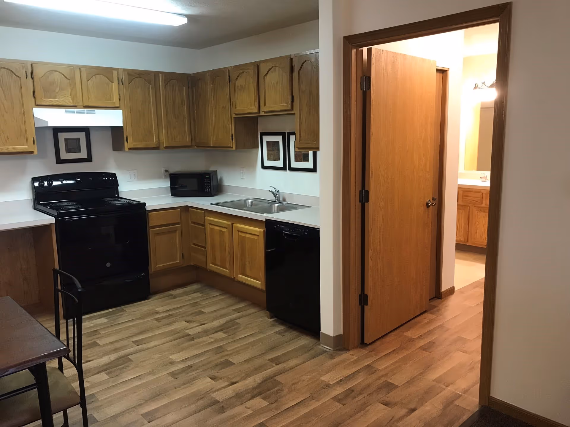 Small kitchen with oak cabinets, black stove and dishwasher, sink and microwave, and an open doorway leading to a hallway/bathroom.