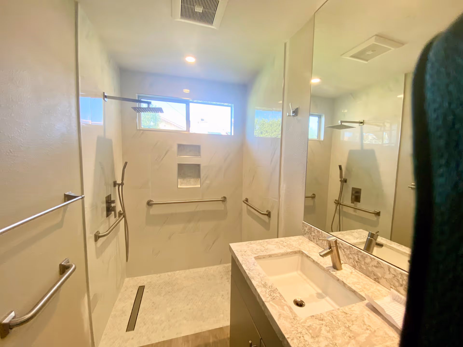 Modern bathroom with a walk-in shower featuring a rainfall showerhead, handheld shower, grab bars, and built-in wall niches. The bathroom has a large mirror above a marble countertop sink and a window letting in natural light.