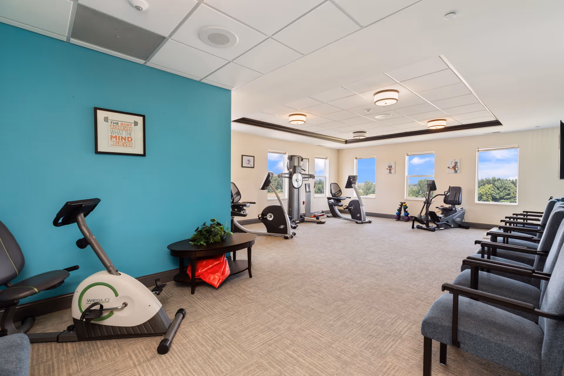 Bright fitness room with exercise bikes, a weight machine, a row of chairs, and a teal accent wall with windows.