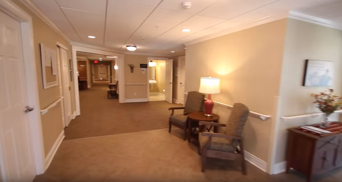 Interior hallway of a senior living facility with beige walls and carpeted floor. Two upholstered chairs with wooden arms are placed around a small round table with a lamp on it. The hallway extends into the distance with doors on either side and a small table with flowers on the right side.