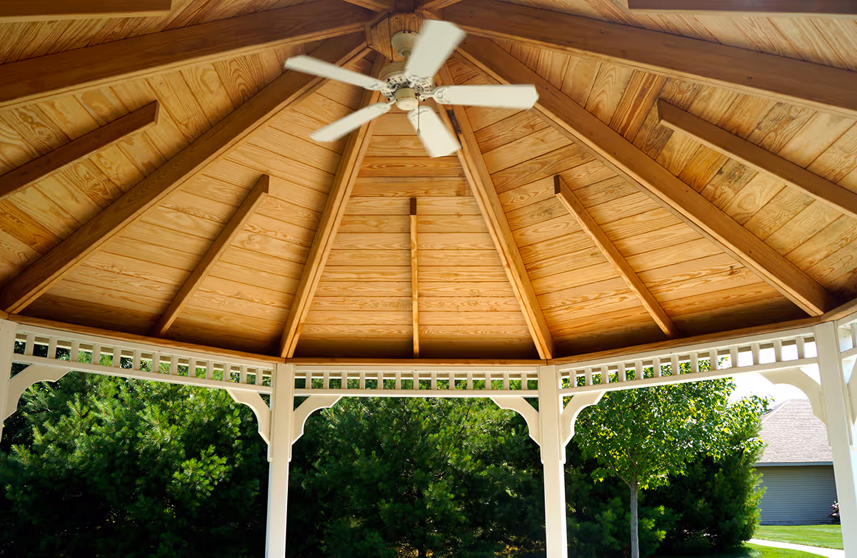 View of the wooden ceiling of a gazebo with a white ceiling fan in the center, surrounded by white support beams and green trees outside.