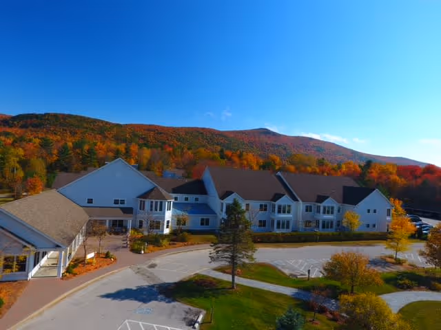 Aerial view of a senior living building with a circular driveway, landscaped grounds, and colorful autumn trees and hills in the background.