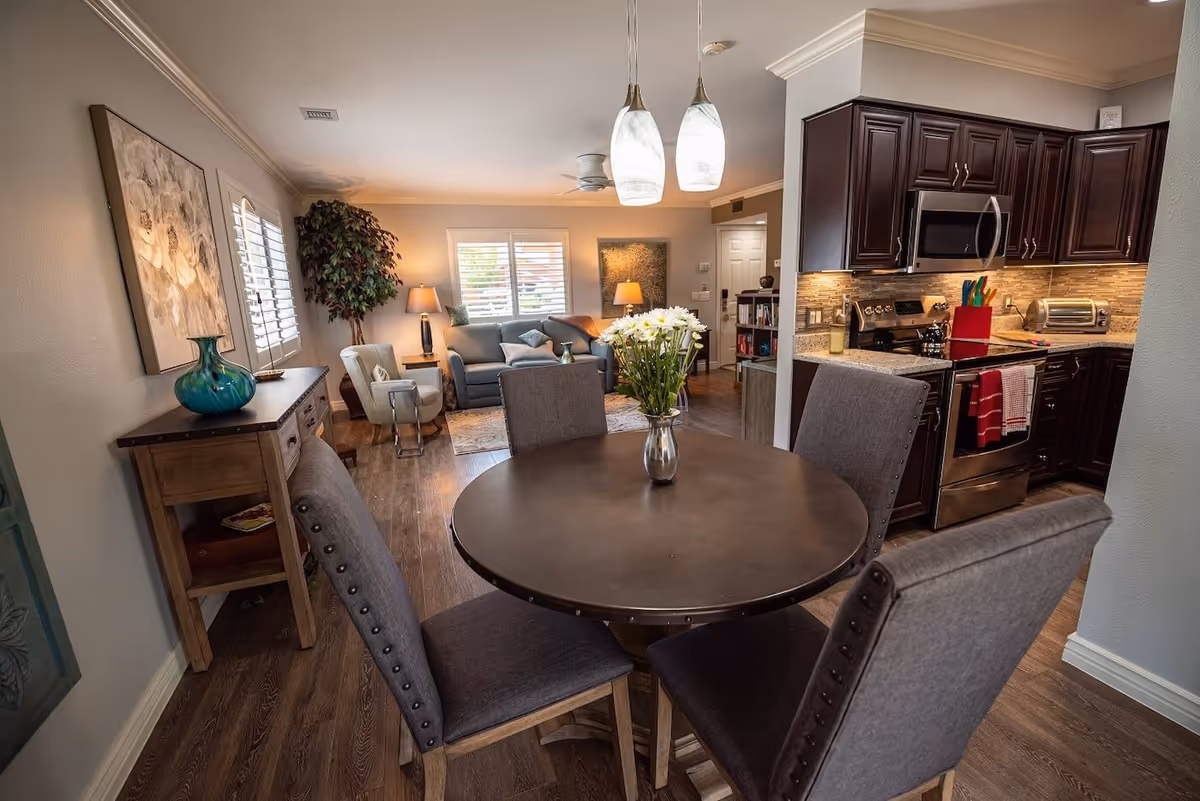 A cozy and well-lit interior space featuring a round dining table with four gray upholstered chairs and a vase of white flowers in the center. In the background, there is a living area with a sofa, armchair, two table lamps, a large potted plant, and wall art. To the right, a modern kitchen with dark wood cabinets, stainless steel appliances, and a countertop with a knife block and toaster is visible.