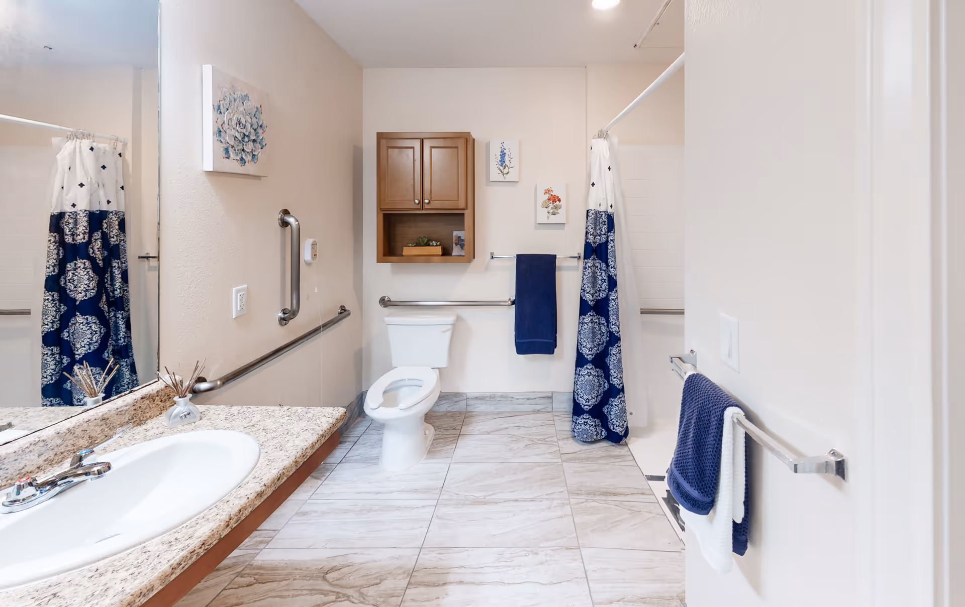 A clean and spacious bathroom with a white toilet, a granite countertop sink, and a shower area with a blue and white patterned curtain. The walls are light-colored with a wooden cabinet above the toilet, two small framed floral artworks, and blue towels hanging on metal bars. The floor is tiled with light-colored tiles.