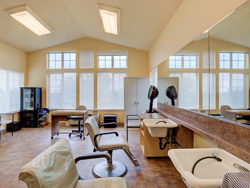 Well-lit salon-style room with styling chairs, shampoo sinks, hooded dryers, and large windows.