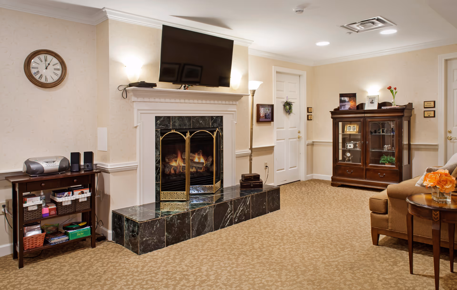 A cozy living room area with a lit fireplace surrounded by black marble tiles and a brass screen. Above the fireplace is a mounted flat-screen TV. To the left, there is a small wooden table with a radio and speakers, along with baskets and books on the shelves below. On the right side, there is a wooden cabinet displaying decorative items and framed pictures. A beige couch with orange flowers on a side table is partially visible. The walls are light-colored with subtle patterns, and there is a clock on the wall showing the time as 11:55. Two white doors and a floor lamp are also visible in the room.