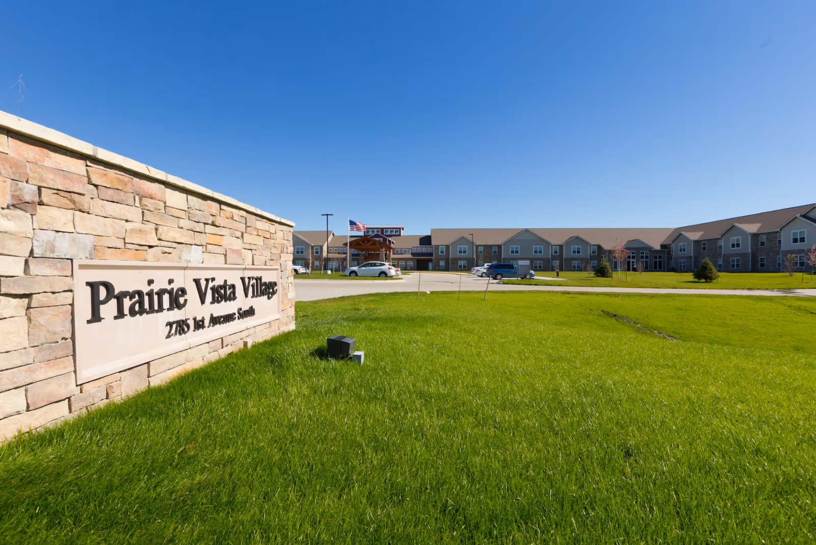 Exterior view of Prairie Vista Village senior living facility with a stone sign in the foreground and a large building with multiple windows and a parking area in the background under a clear blue sky.