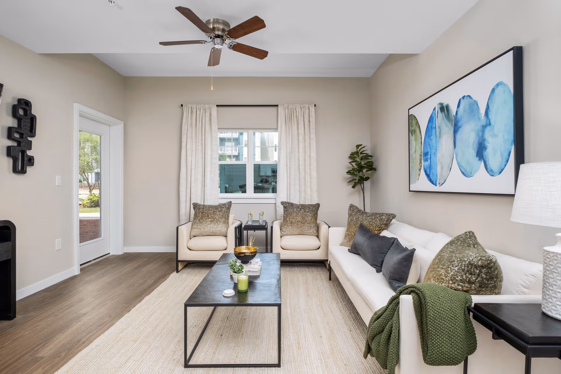 A modern living room with a white sofa adorned with green and gray pillows and a green throw blanket. Two beige armchairs with matching pillows are placed in front of a window with cream-colored curtains. A black rectangular coffee table with decorative items sits on a light beige rug over wooden flooring. A ceiling fan with wooden blades is mounted on the white ceiling. A large abstract painting with blue and green circular shapes hangs on the right wall, and a small green plant is in the corner.