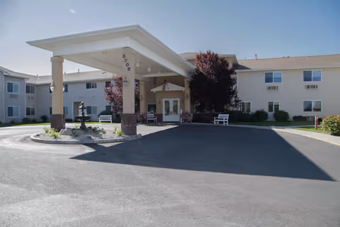 Front exterior view of a two-story senior living facility building with a covered entrance supported by columns, a circular driveway, benches, and landscaping including trees and bushes under a clear blue sky.