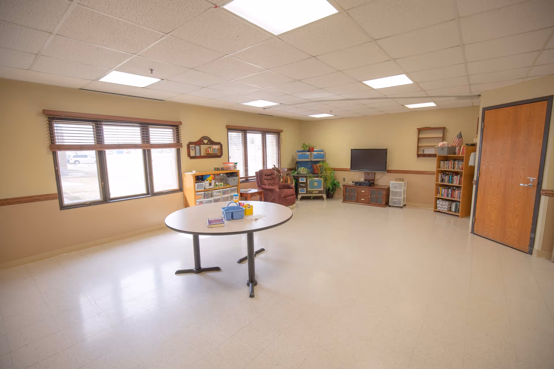 A spacious room with beige walls and a tiled floor, featuring a round table with some items on it, a comfortable recliner chair, a TV mounted on the wall with a cabinet below, bookshelves filled with books, and several windows with wooden blinds letting in natural light.