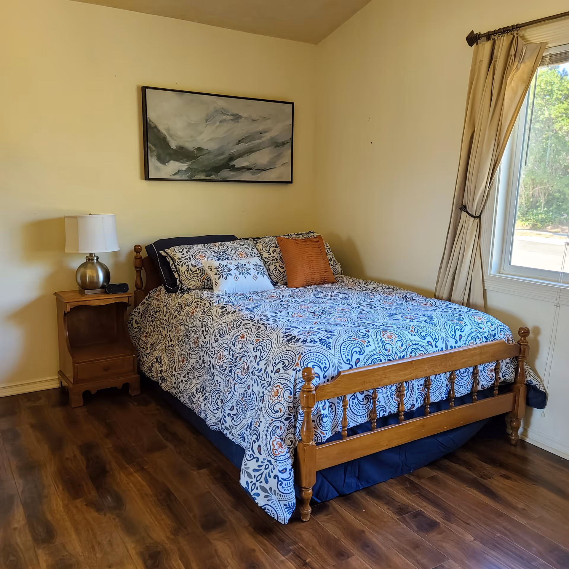 A bedroom with a wooden bed frame and a bed covered with a blue and white patterned comforter and multiple pillows. There is a wooden nightstand with a silver lamp and a small black device on it. A large window with beige curtains is on the right side, letting in natural light. A framed abstract painting hangs on the wall above the bed. The floor is wooden.