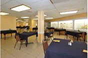 A spacious room with multiple tables covered in dark tablecloths, each surrounded by brown chairs. The room has a tiled floor, fluorescent ceiling lights, and large windows along one wall. The setting appears to be a dining or activity area in an assisted living facility.