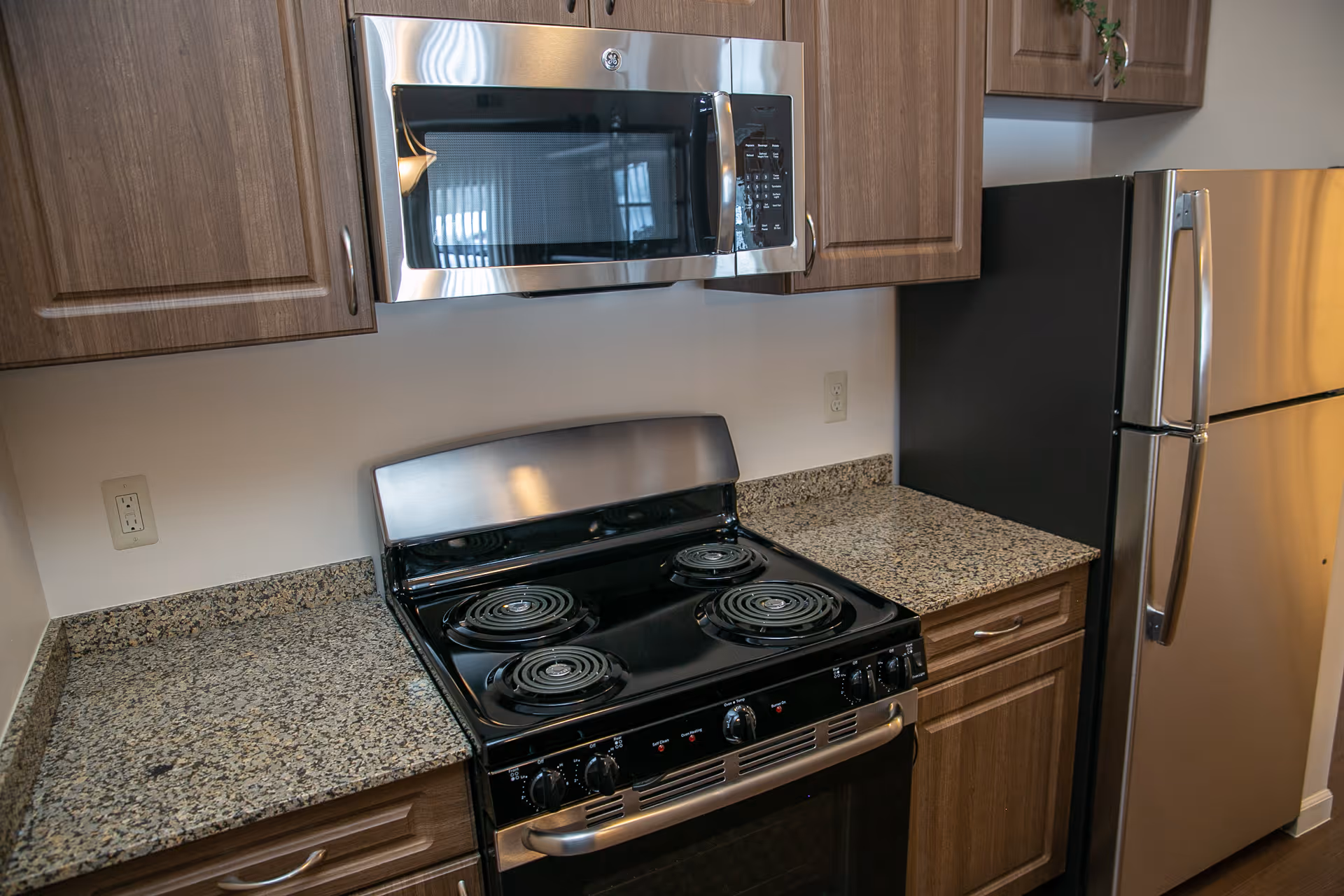 Modern kitchen with an electric stove and oven, over-the-range microwave, granite countertops, wooden cabinets, and a stainless steel refrigerator.