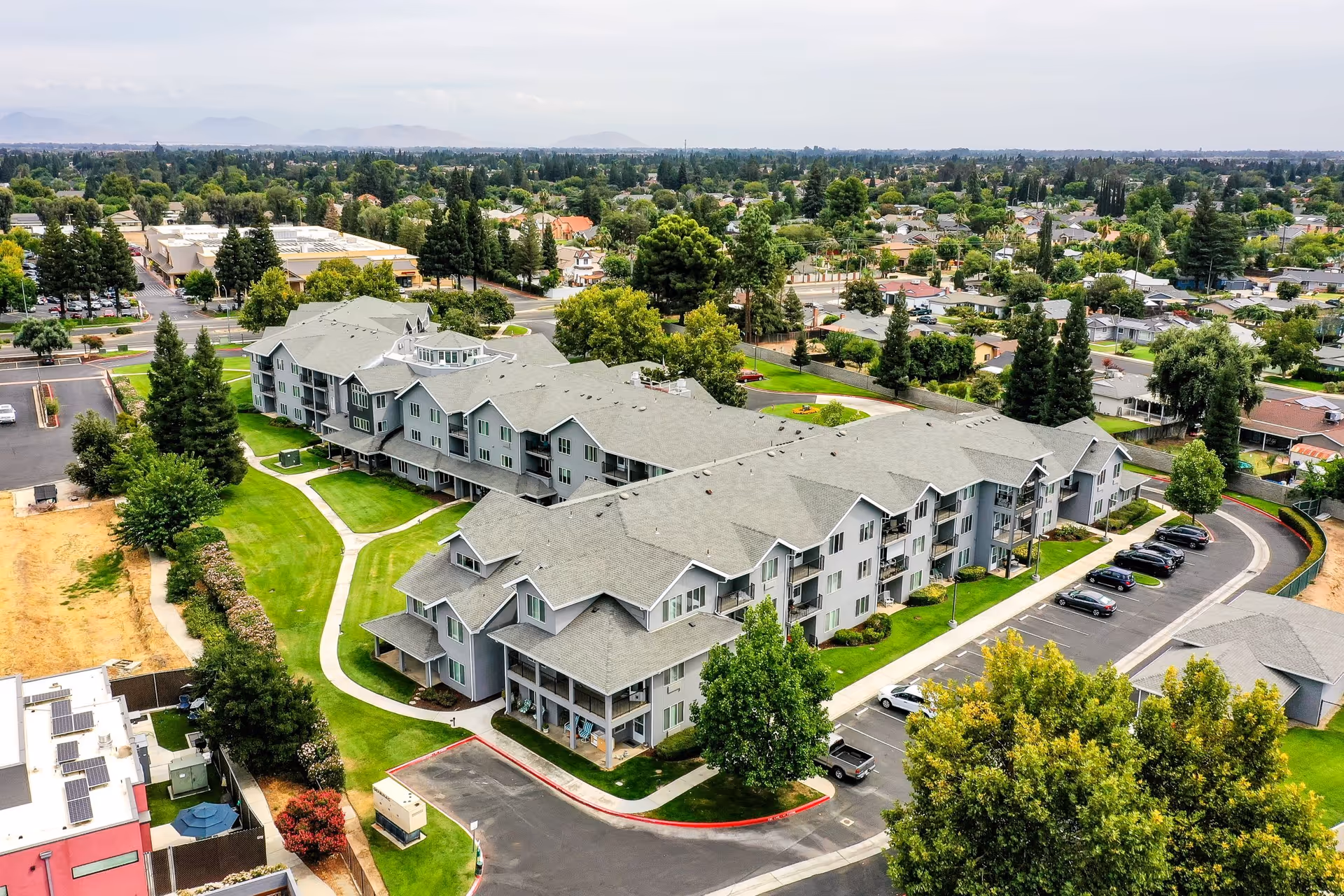 Aerial view of Solstice Senior Living at Clovis, showing a large multi-story residential building with gray roofs surrounded by green lawns, trees, and parking areas. The surrounding neighborhood with houses and streets is visible in the background under a clear sky.