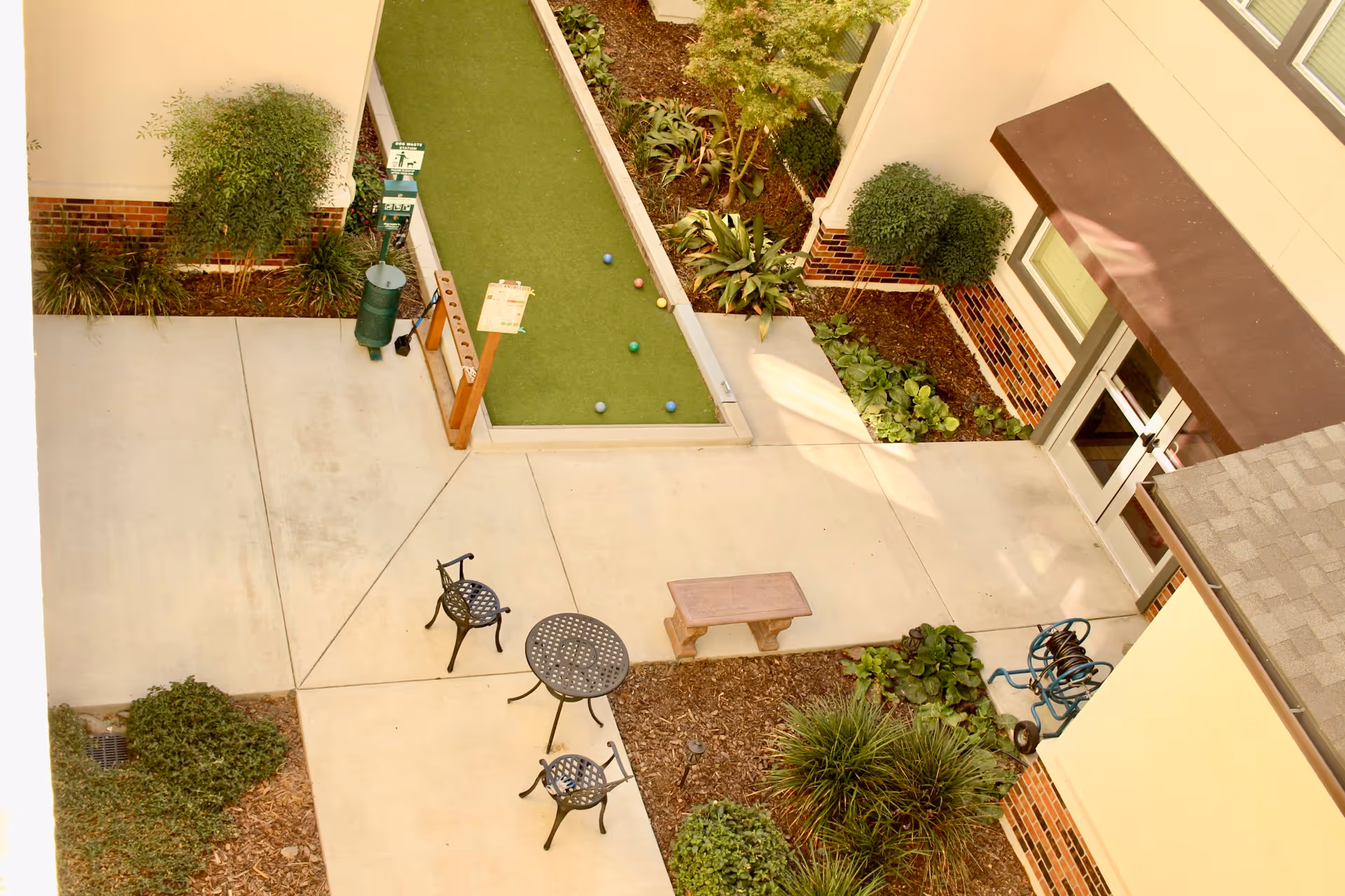 View of an outdoor courtyard area at Summerset Senior Living, Lincoln featuring a bocce ball court with several bocce balls on it, a small round metal table with two matching chairs, a wooden bench, and various plants and shrubs along the edges of the concrete pathways and building walls.