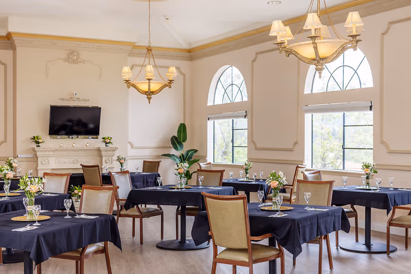 A bright dining room with multiple tables covered in dark tablecloths, each set with glassware, plates, and floral centerpieces. The room features large arched windows letting in natural light, elegant chandeliers hanging from the ceiling, and a wall-mounted TV above a decorative fireplace.