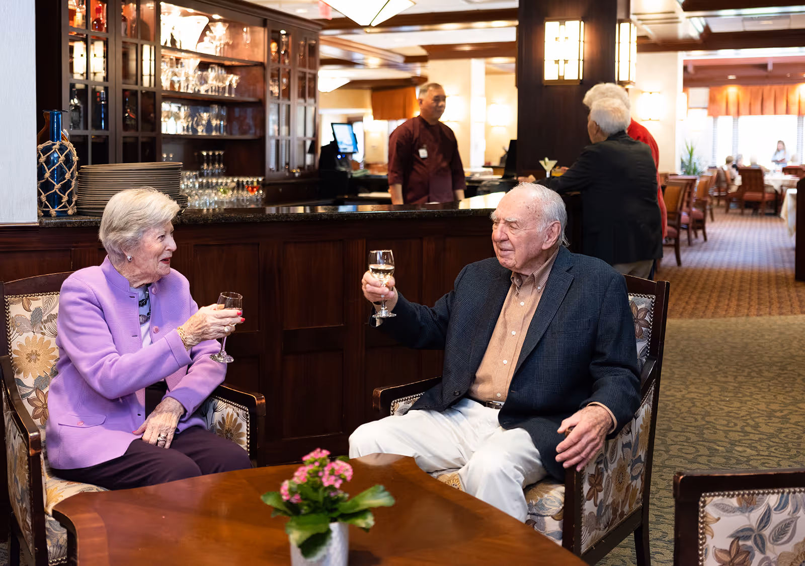 Two elderly people sitting in upholstered chairs at a wooden table with a small potted plant, raising glasses in a toast inside a senior living community lounge area with a bar and other residents in the background.