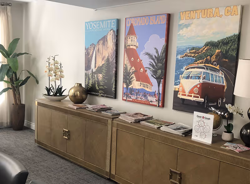 Interior view of a senior living facility room with a long beige cabinet against the wall. On top of the cabinet are several stacks of magazines, decorative vases, and a small sign. Above the cabinet, three colorful travel posters are displayed on the wall, depicting Yosemite National Park, Coronado Island, and Ventura, CA. A tall potted plant is placed in the corner near a window with sheer curtains.