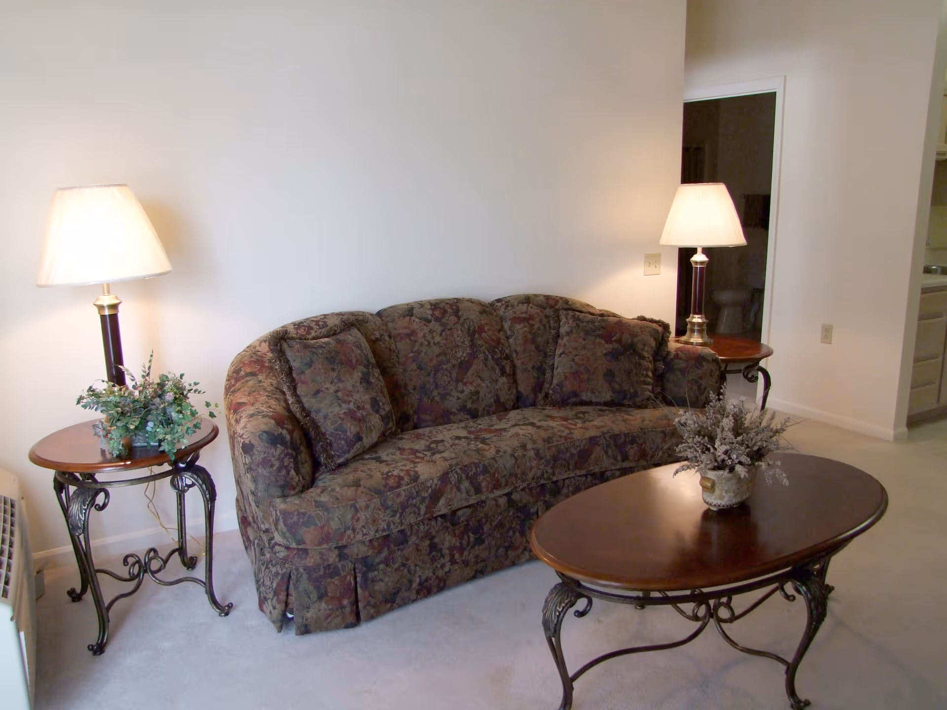A living room with a floral patterned sofa, two matching side tables each with a lamp and a small plant, and a wooden coffee table with a floral arrangement. The walls are plain white and there is an open doorway leading to another room.