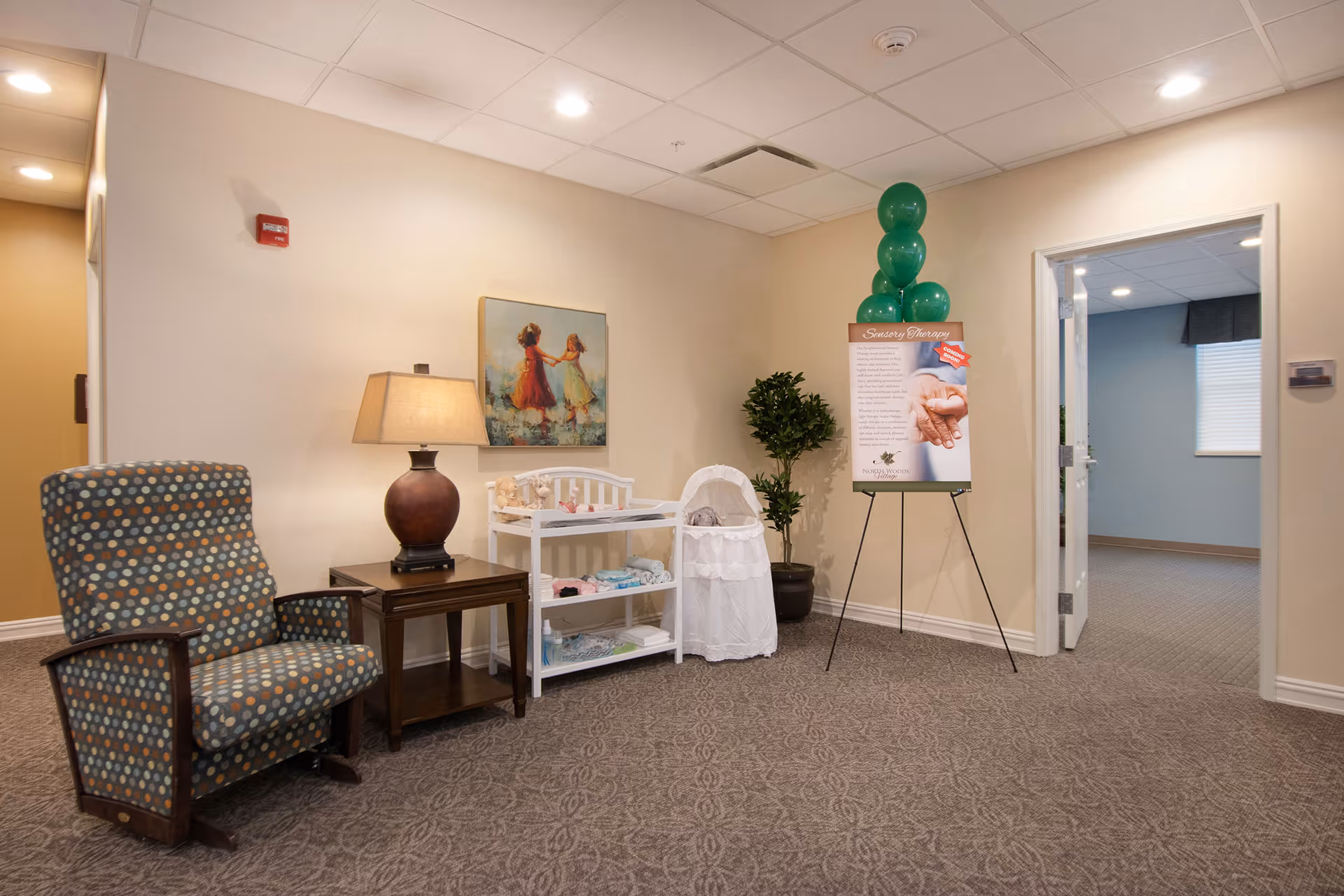 A cozy corner in a senior living facility featuring a patterned armchair, a wooden side table with a lamp, a white changing table with baby items, a bassinet, a potted plant, and a stand with a sign and green balloons. The walls are beige and there is an open doorway leading to another room with blue walls and a window.