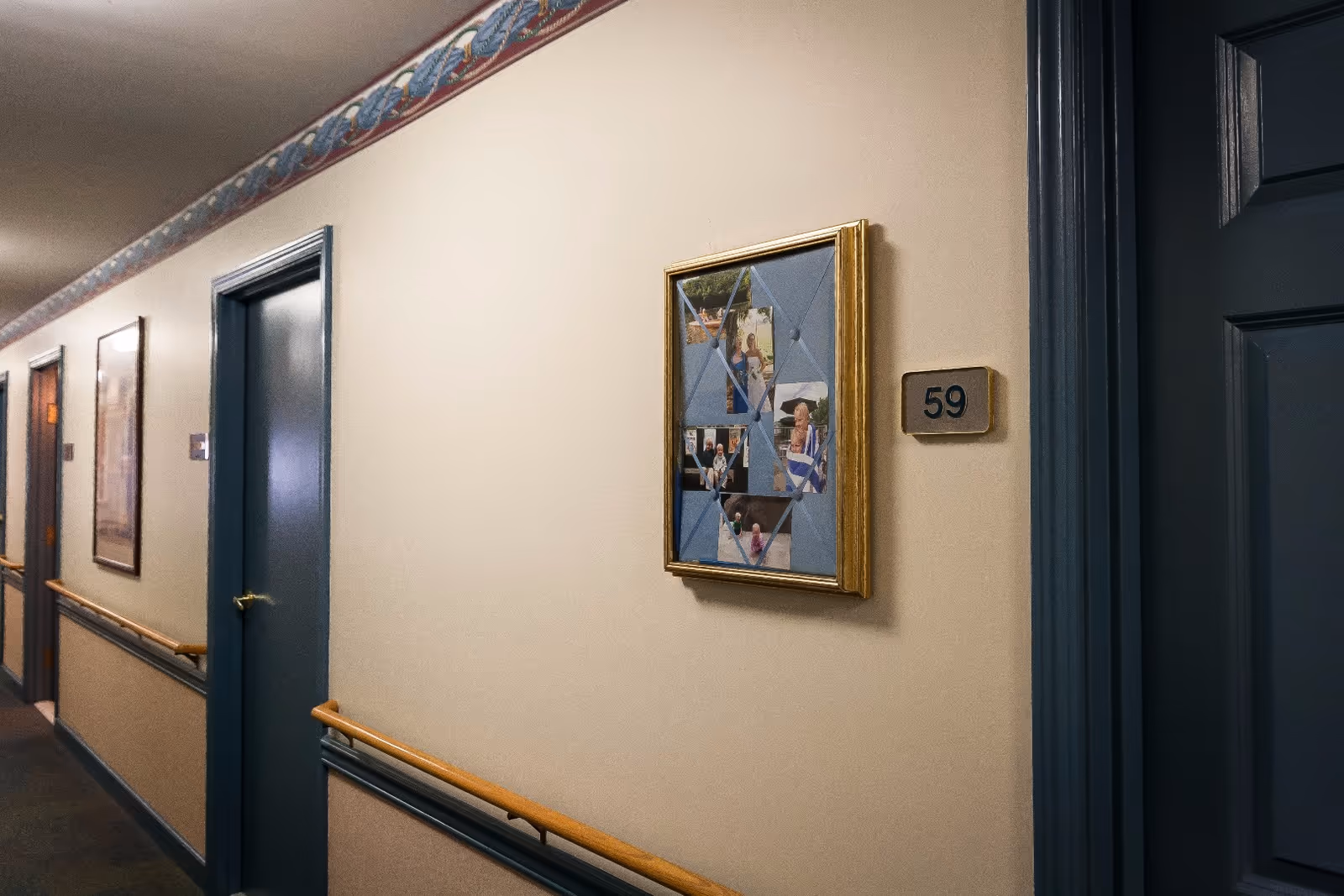 A hallway in a senior living facility with beige walls and blue doors. There is a framed collage of photos on the wall next to a door labeled with the number 59. The hallway has a wooden handrail and decorative border near the ceiling.