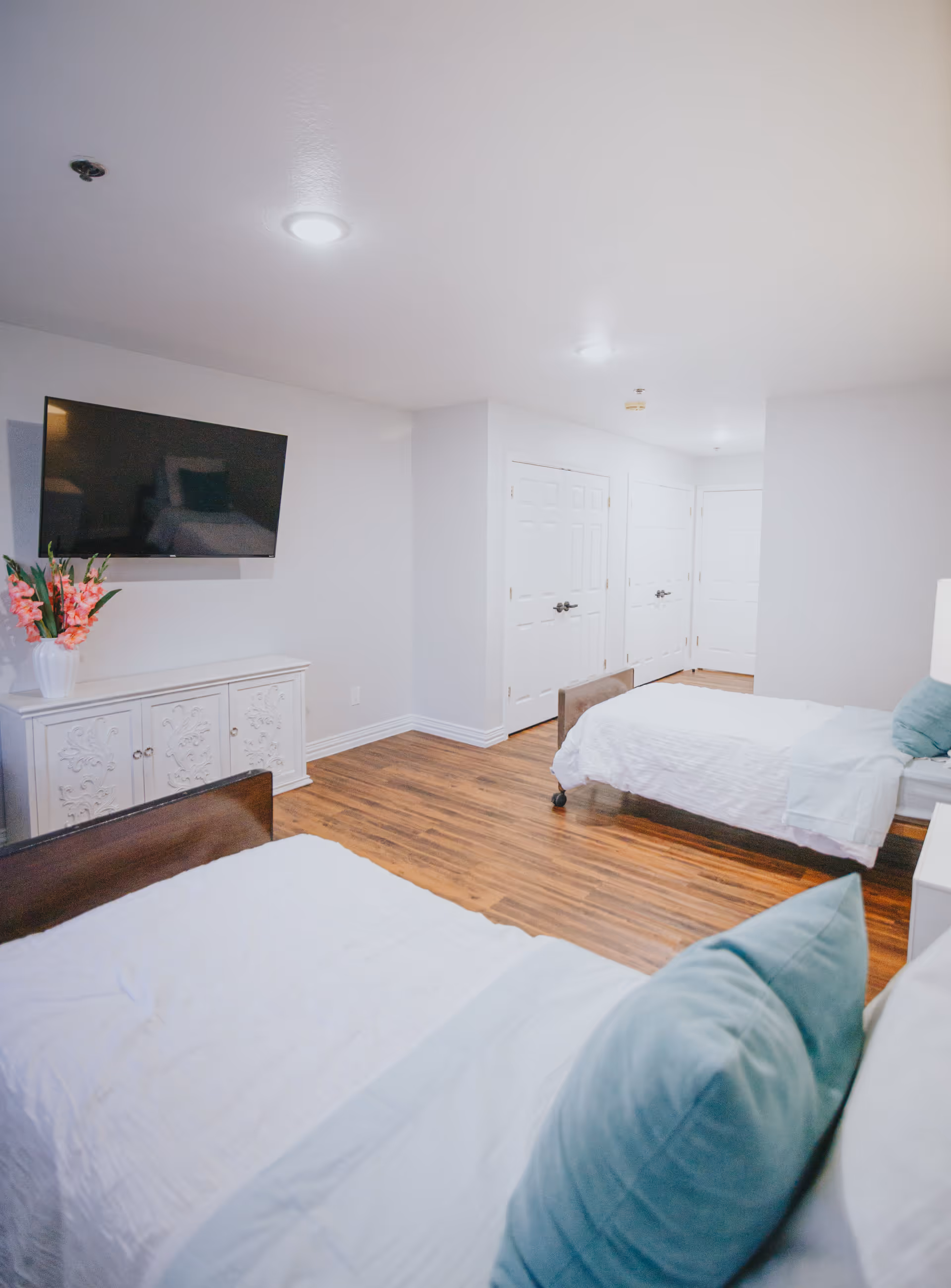 A bright bedroom with two single beds, each with white bedding and light blue pillows. There is a white cabinet with floral designs on the doors, a vase with pink flowers on top, and a flat-screen TV mounted on the wall above the cabinet. The room has wooden flooring and white walls with multiple white doors in the background.
