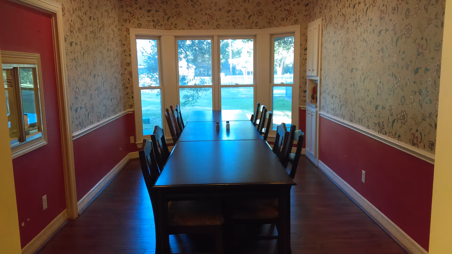 Long rectangular dining table with multiple chairs in a narrow dining room facing a large bay window overlooking a grassy yard.