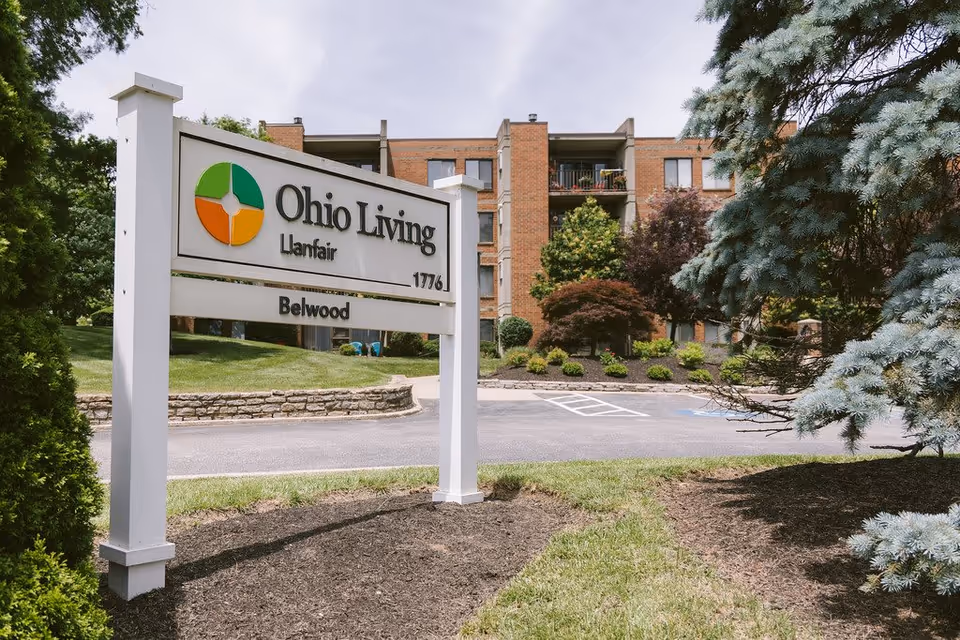 Outdoor view of Ohio Living Llanfair facility sign with a brick building and landscaped greenery in the background under a partly cloudy sky.