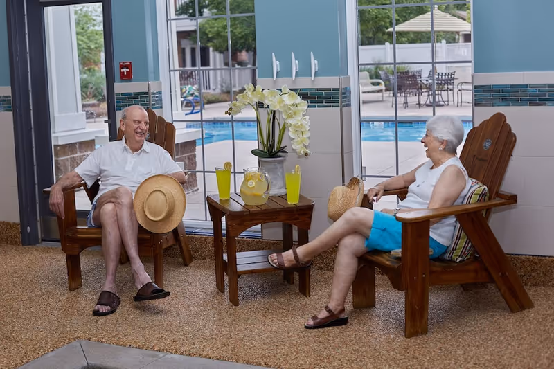 An elderly man and woman sitting on wooden chairs indoors near large windows overlooking a swimming pool. They are smiling and appear to be enjoying a conversation. A small wooden table between them holds a pitcher of lemonade, two glasses with lemon slices, and a vase with white orchids. Both are holding straw hats.