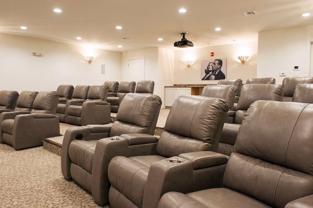A cozy theater room with multiple rows of comfortable gray recliner chairs arranged in tiers, a ceiling-mounted projector, soft wall sconces, and a black and white framed picture on the back wall.
