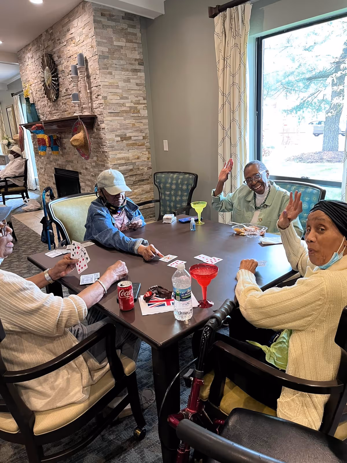 Four elderly individuals sitting around a square table playing cards in a well-lit room with a large window. The room has a stone fireplace with decorative items on the mantel and patterned curtains. Drinks, including a can of Coca-Cola, a water bottle, and colorful margarita glasses, are on the table. Two of the individuals are smiling and waving at the camera.