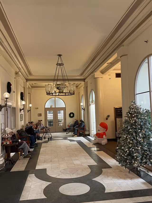 Spacious decorated lobby with a chandelier, seating along the walls where several seniors sit with walkers, and a Christmas tree and holiday decorations.