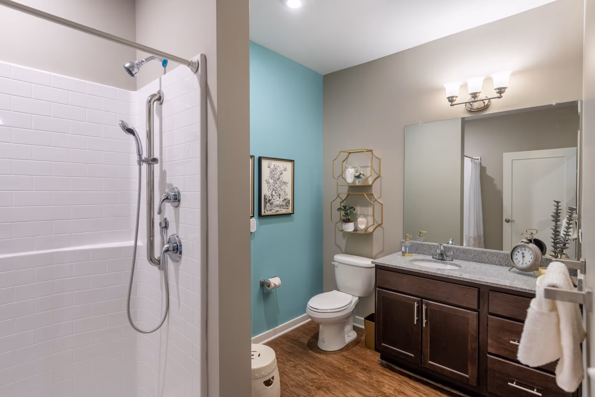 A modern bathroom featuring a white tiled shower with two showerheads and a grab bar, a toilet against a teal accent wall, a wooden vanity with a granite countertop, a large mirror, and decorative shelving above the toilet. The floor is wood-style, and the lighting fixture above the mirror has three lights.