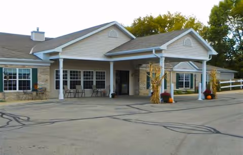 Exterior view of a single-story assisted living facility building with a covered entrance supported by white columns. The building has beige siding, stone accents, and several windows. There are autumn decorations including cornstalks and pumpkins near the entrance. Trees with green foliage are visible in the background.