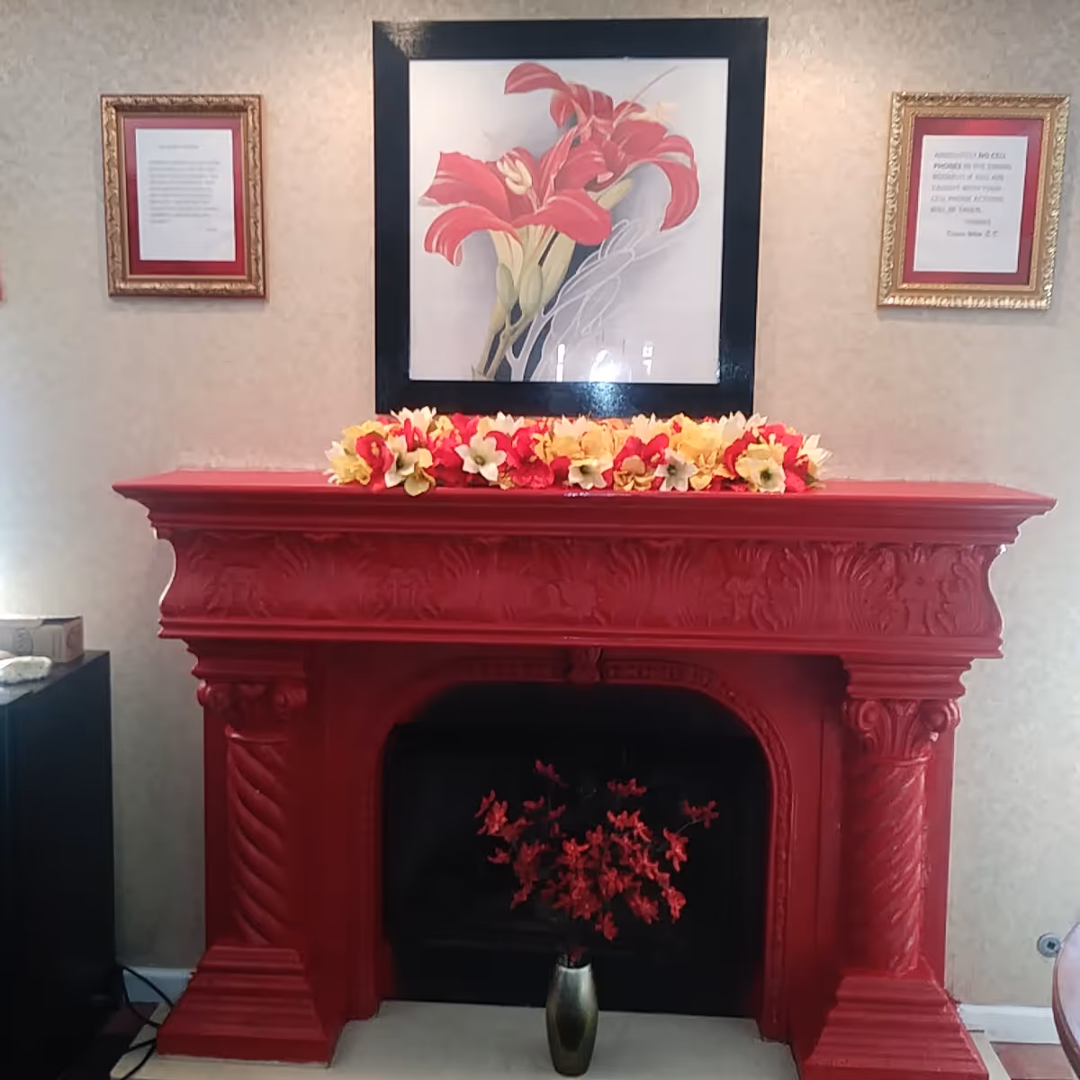 Red ornamental fireplace mantel with flowers on top and a framed floral picture above.